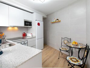 Una cuina moderna amb armaris blancs, microones, nevera vermella, rentapelats i una petita zona de menjador amb taula i cadires.