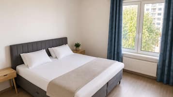 A bedroom with a bed, a nightstand, a potted plant, a window with curtains, and a radiator.