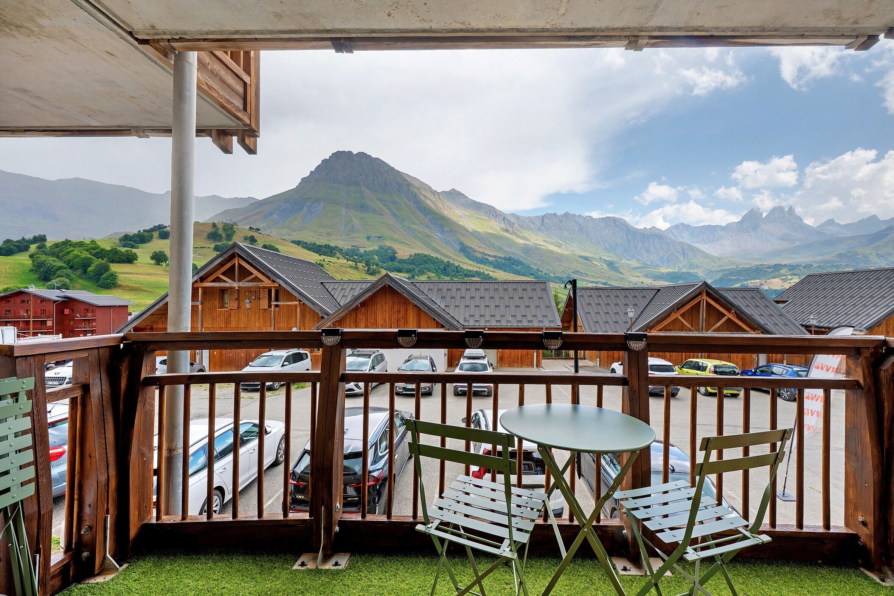 Un balcon en bois offrant une vue sur une chaîne de montagnes, un espace de stationnement et une table avec des chaises.