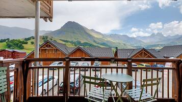 Un balcon en bois offrant une vue sur une chaîne de montagnes, un espace de stationnement et une table avec des chaises.