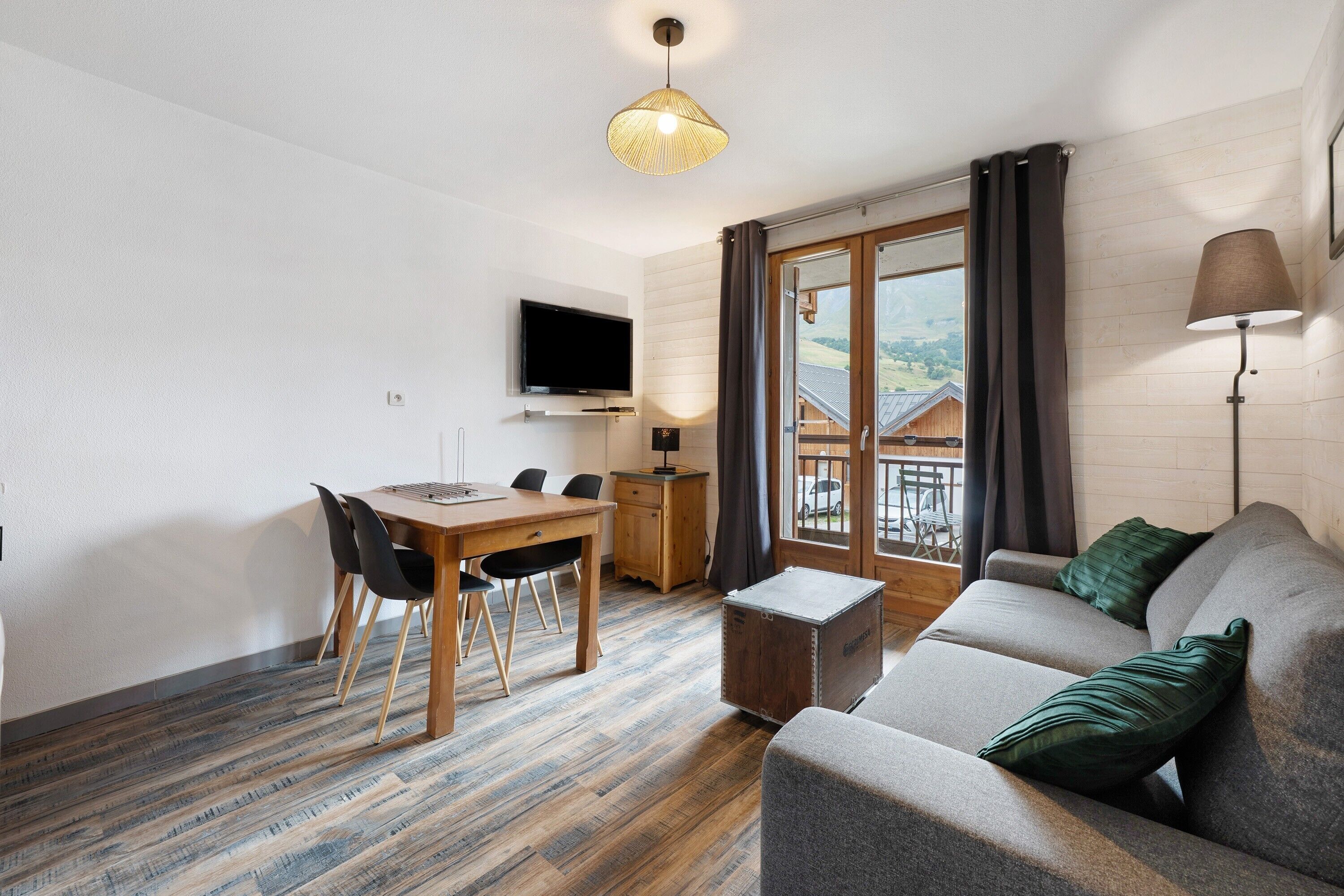 Een moderne woonkamer met een houten eettafel, zwarte stoelen, een grijze bank met groene kussens, een aan de muur bevestigde televisie en een raam met gordijnen.