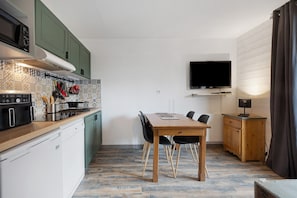 Een kleine keuken met een magnetron, fornuis en diverse keukengerei. Een eettafel met stoelen staat in het midden, en een flatscreen televisie is aan de muur bevestigd.