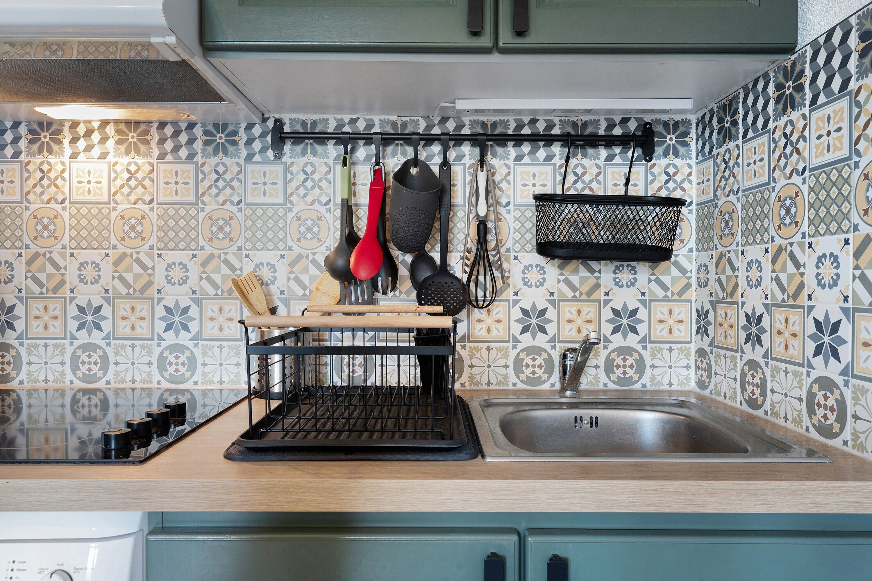 Een keuken met een betegelde spatwand, een hangrek met keukengerei, een spoelbak en een fornuis.