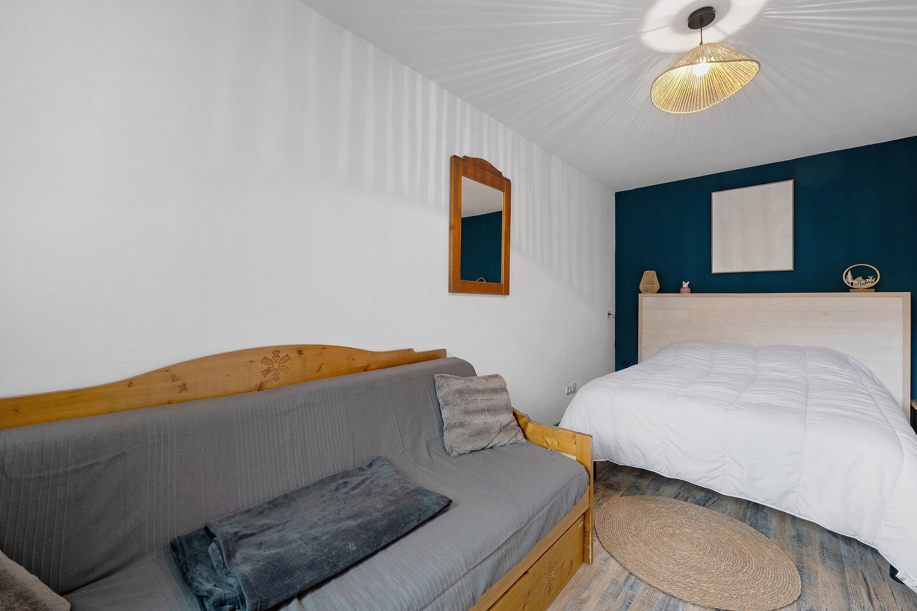 Une chambre avec un lit doté d’une tête de lit en bois, un canapé gris avec un plaid, un tapis rond tissé et un luminaire suspendu au plafond.