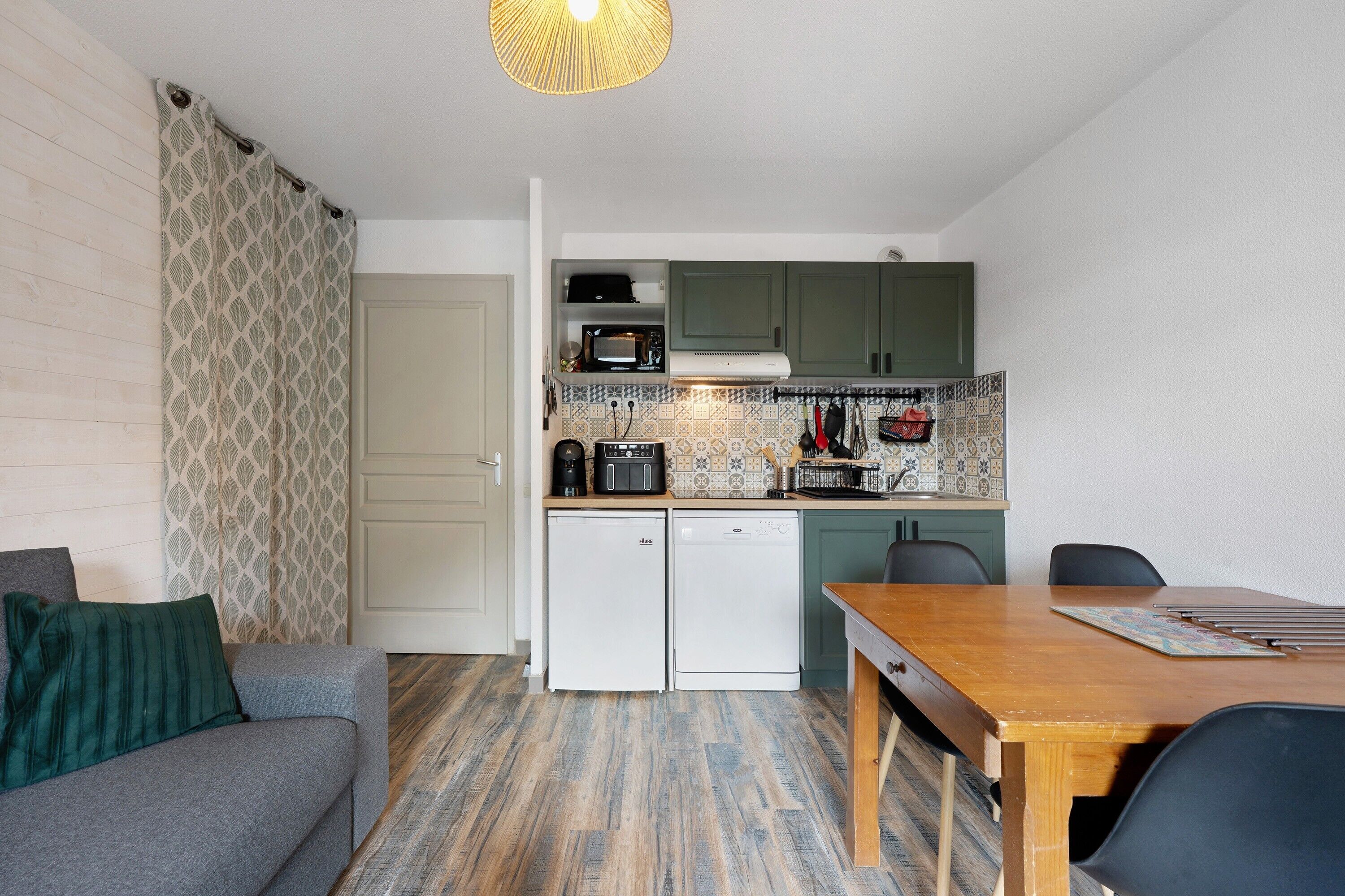 Une cuisine compacte équipée d’un four à micro-ondes, d’une machine à café et d’un petit réfrigérateur. On y trouve une table à manger avec des chaises, ainsi qu’un canapé agrémenté d’un plaid à motifs.
