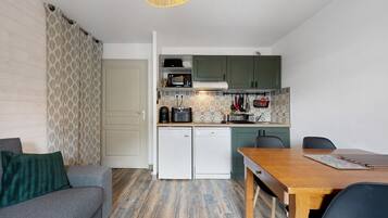 Une cuisine compacte équipée d’un four à micro-ondes, d’une machine à café et d’un petit réfrigérateur. On y trouve une table à manger avec des chaises, ainsi qu’un canapé agrémenté d’un plaid à motifs.