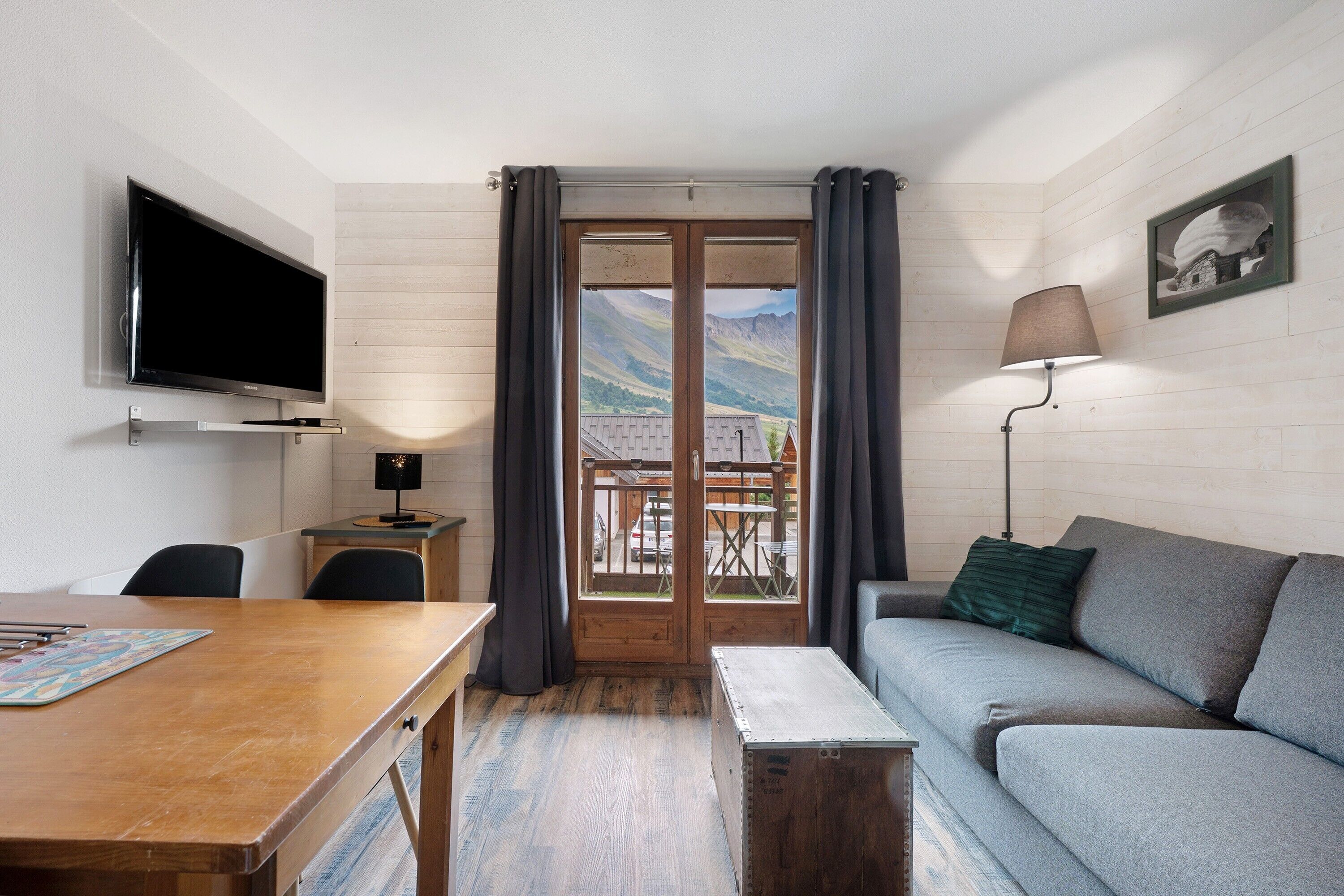 Een kamer met televisie, een tafel, een bank, een lamp en uitzicht op bergen door een deur.