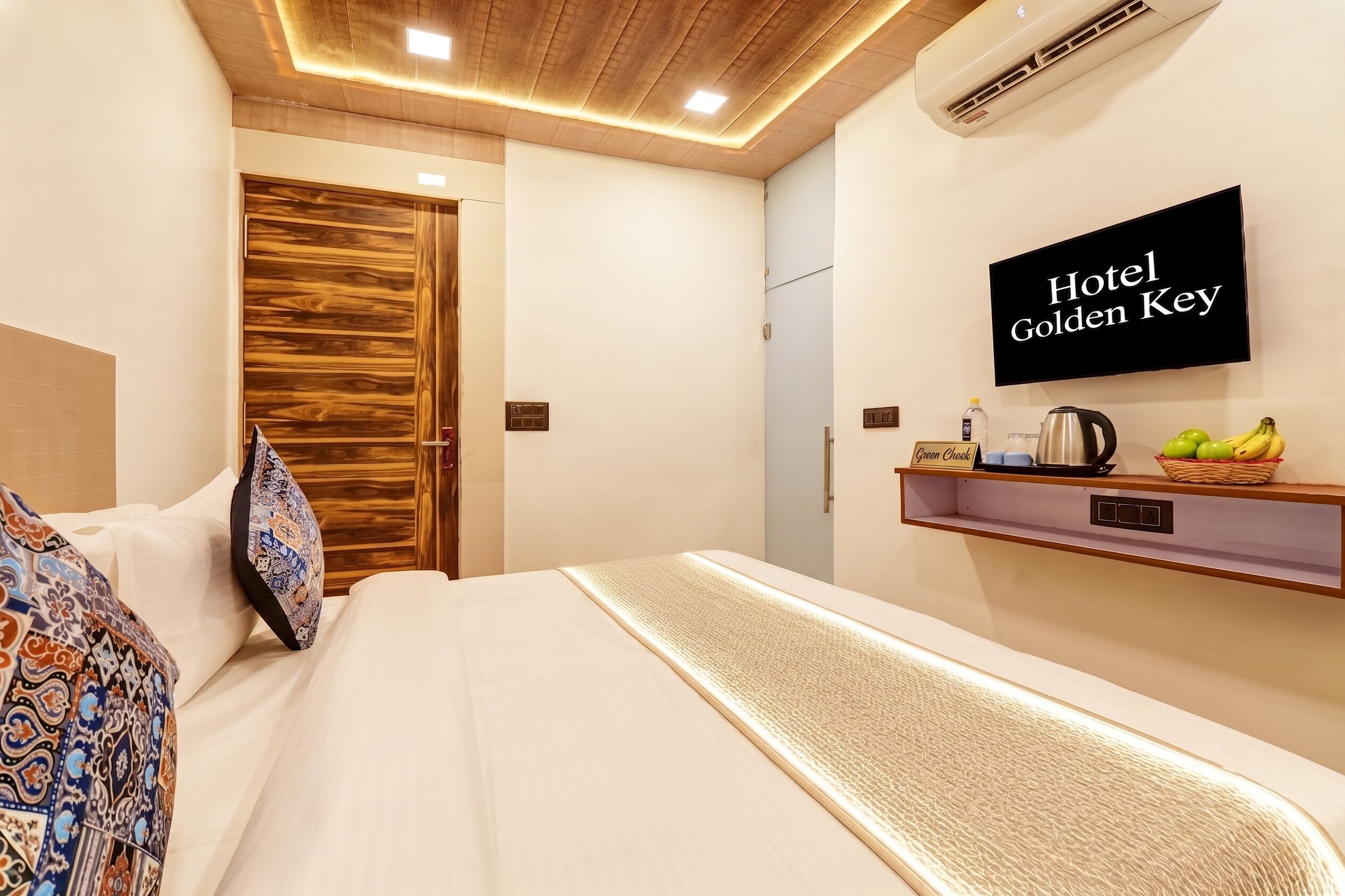 A hotel room with a bed, a wooden door, a wall-mounted TV, and a shelf with a kettle and fruits.