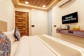 A hotel room with a bed, a wooden door, a wall-mounted TV, and a shelf with a kettle and fruits.