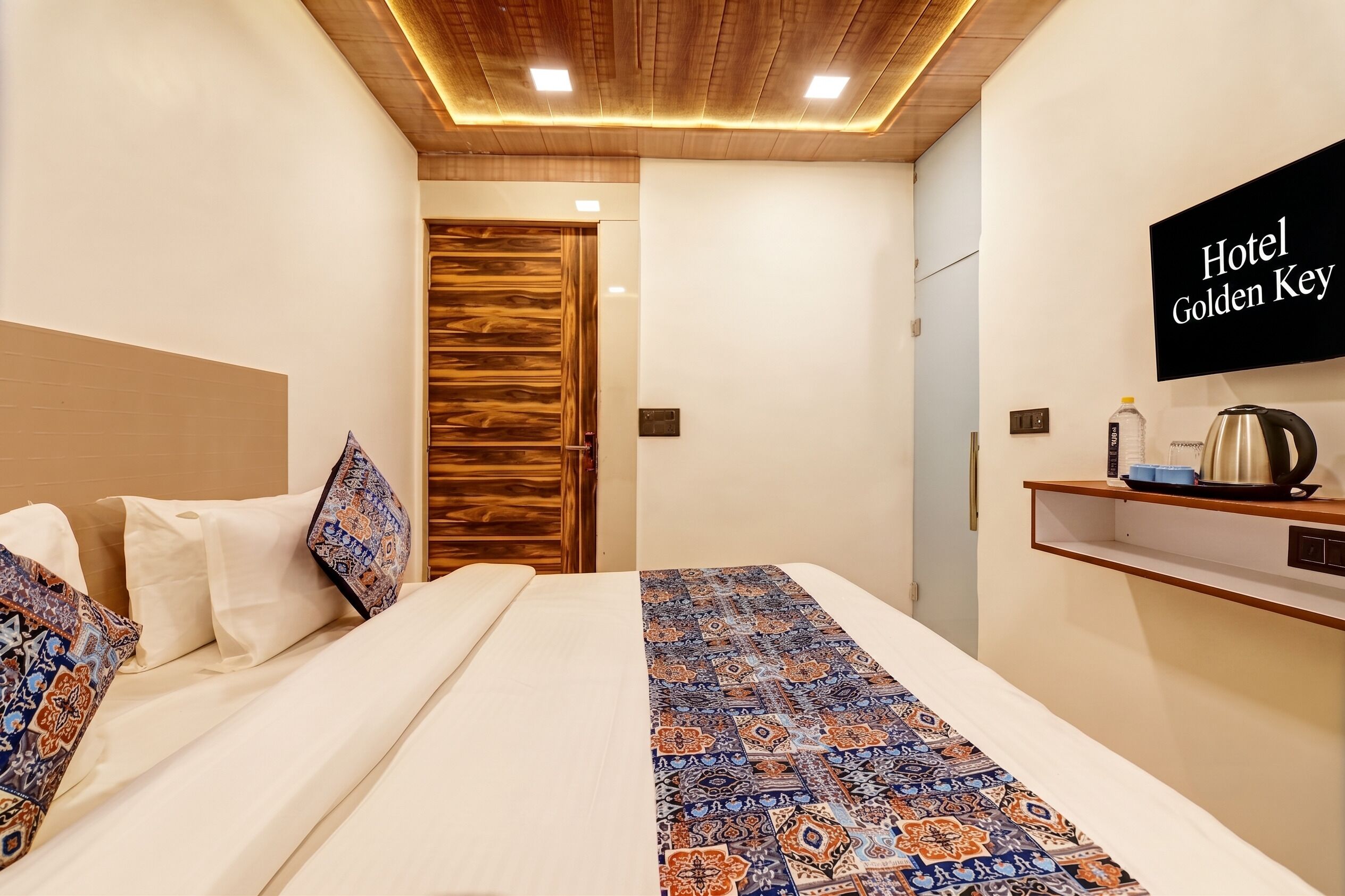 Habitació d'hotel amb un llit, un panell de fusta al capçal, un correll de llit estampat, un televisor penjat a la paret i una estant amb una tetera i tasses.