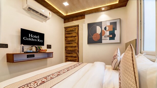 A hotel room with a bed, a flat-screen TV displaying 'Hotel Golden Key', a wall-mounted shelf with amenities, and a modern abstract painting.