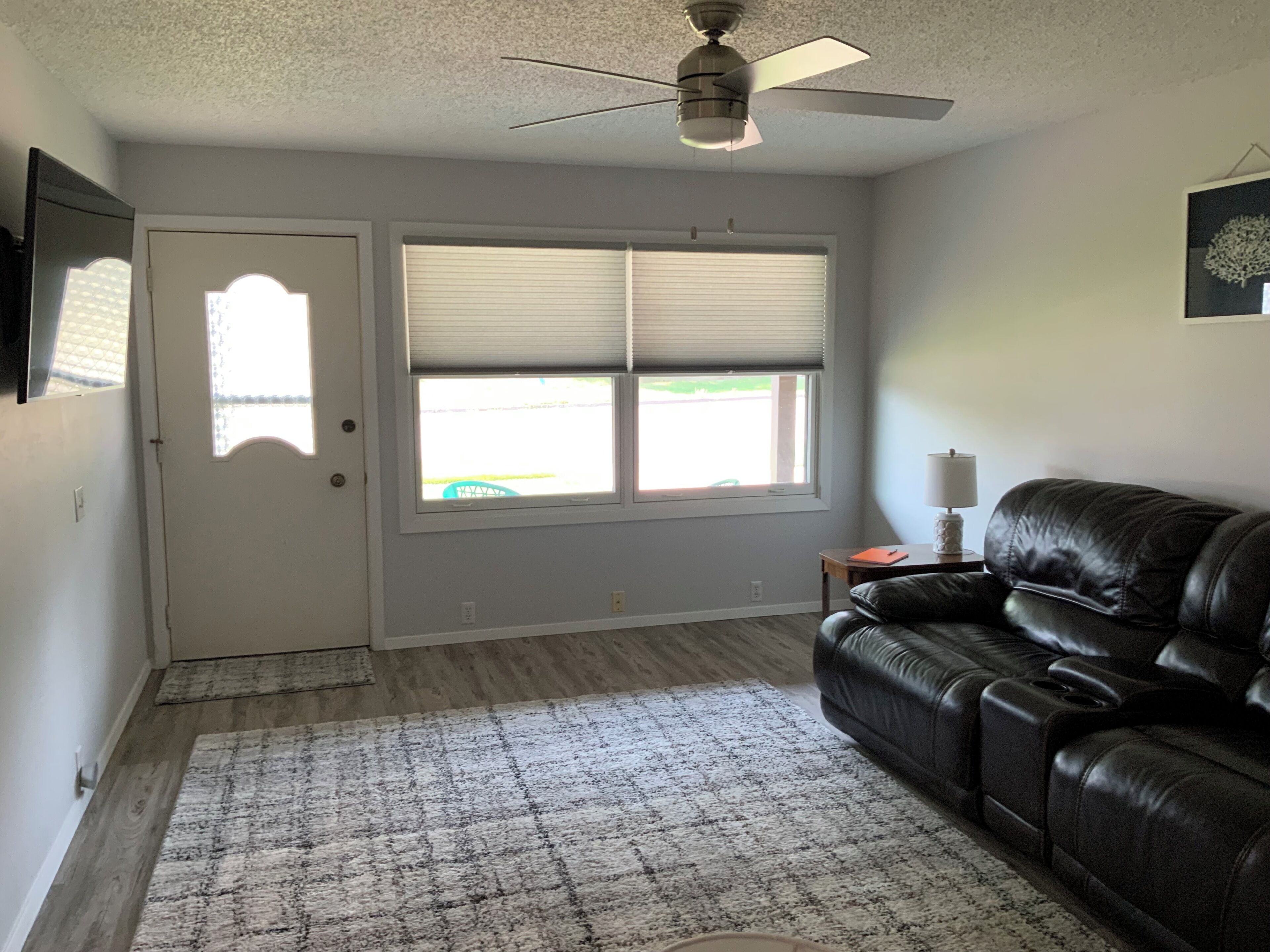 A living room with a black leather sofa, a ceiling fan, a window with blinds, and a door.