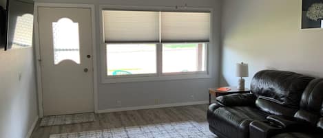 Ein Wohnzimmer mit schwarzem Ledersofa, Deckenventilator, einem Fenster mit Jalousien und einer Tür.
