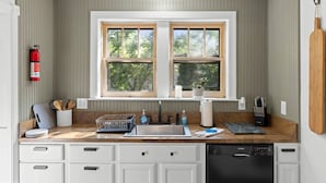 A kitchen with a double sink, wooden countertop, white cabinets, and a window with a view of trees.