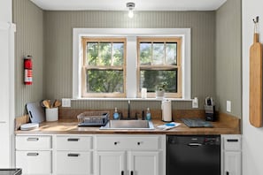 A kitchen with a double sink, wooden countertop, white cabinets, and a window with a view of trees.
