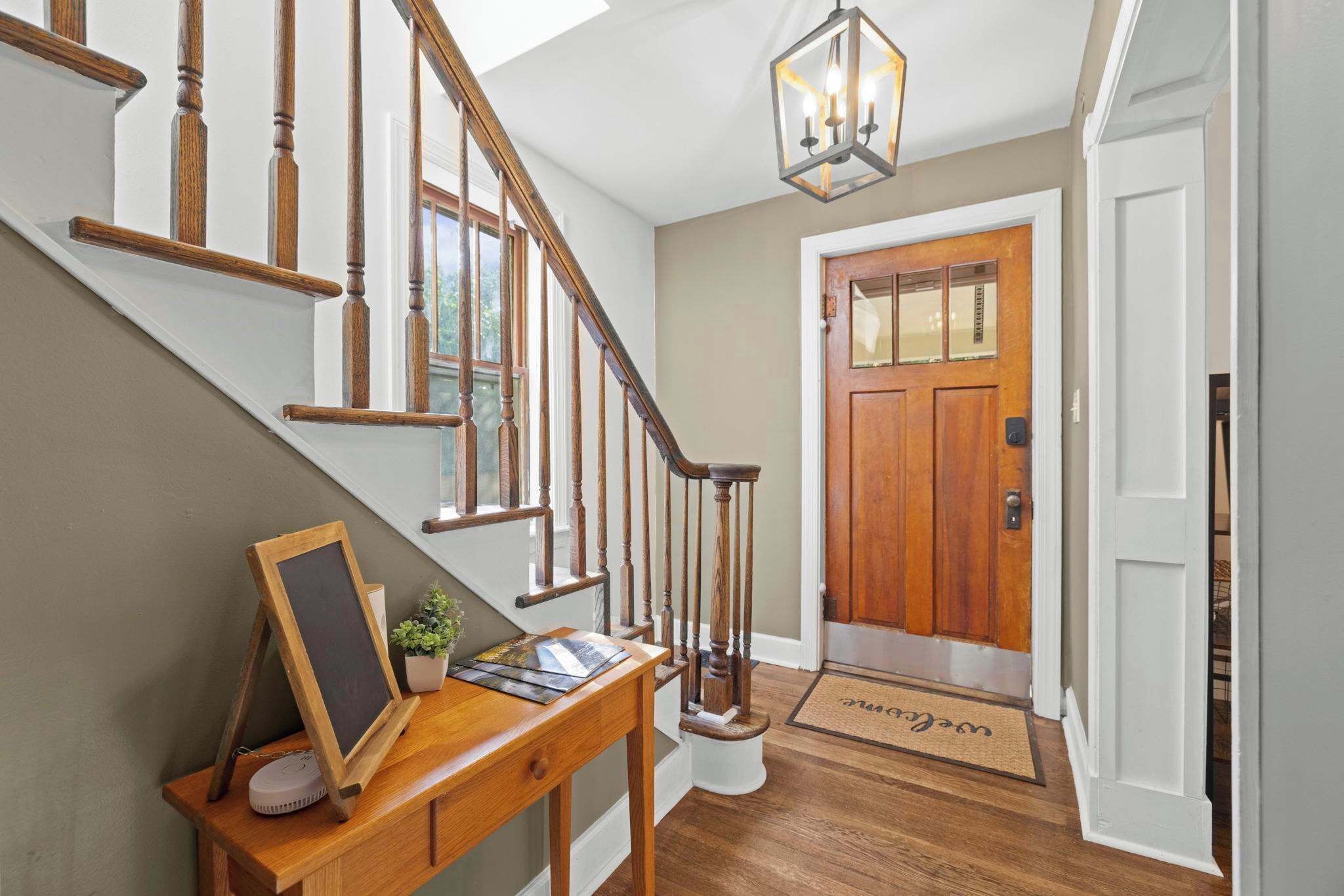 A wooden staircase with a handrail, a desk with a chalkboard, a potted plant, and a welcome mat.