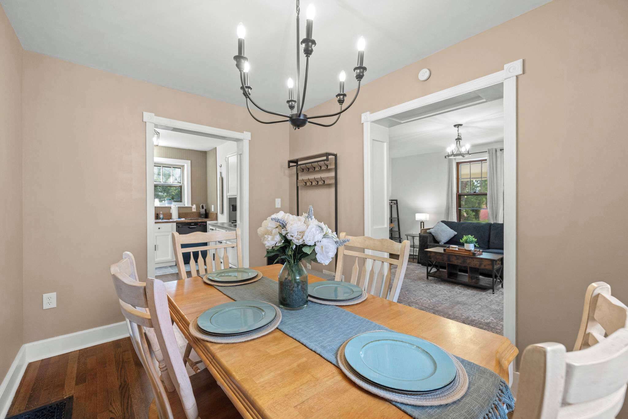 A dining room with a wooden table set for four, a chandelier, and a view into the kitchen and living area.