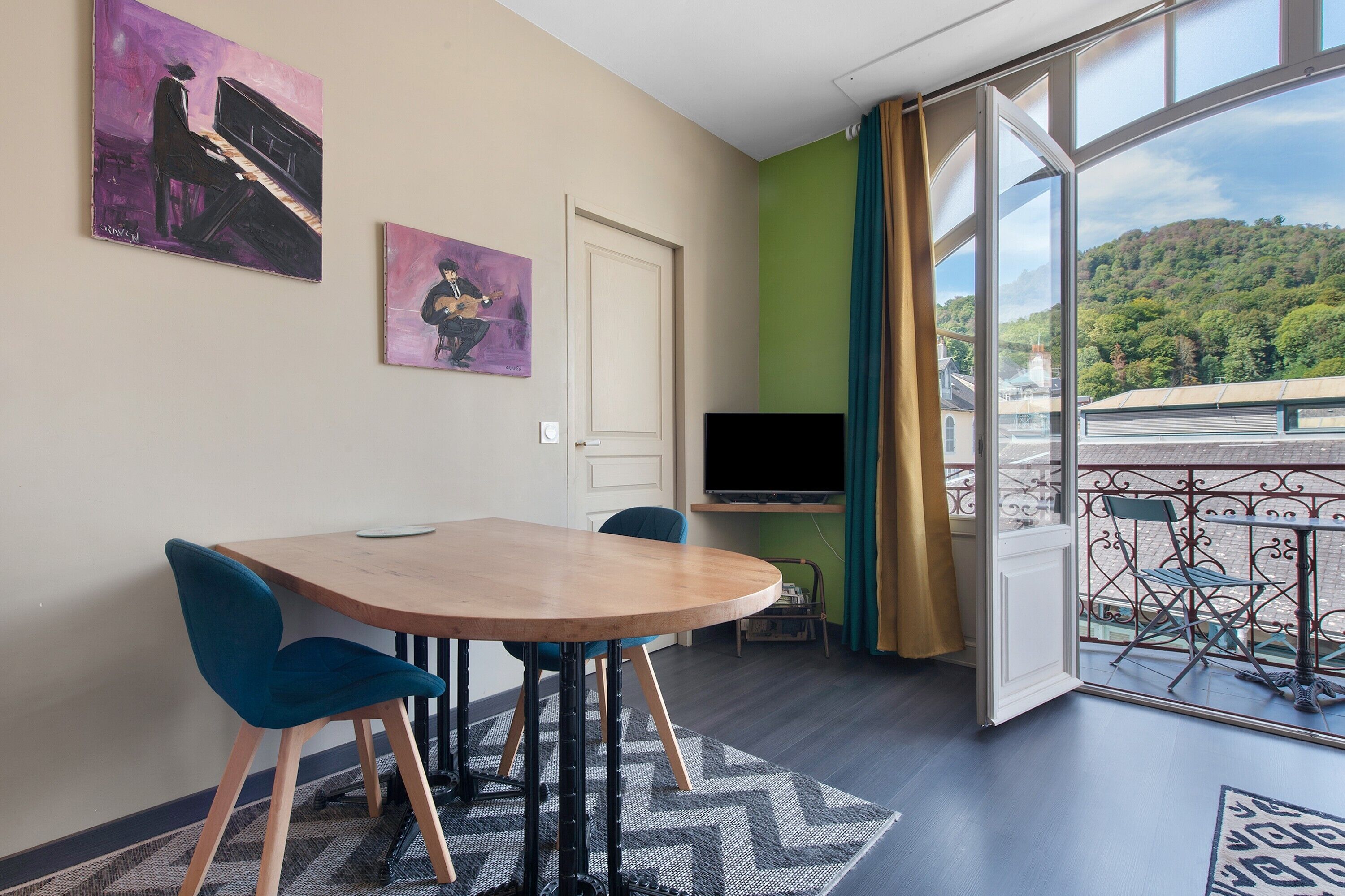 Una habitación con una mesa de comedor, sillas, un televisor y un amplio ventanal con vistas a una colina boscosa.