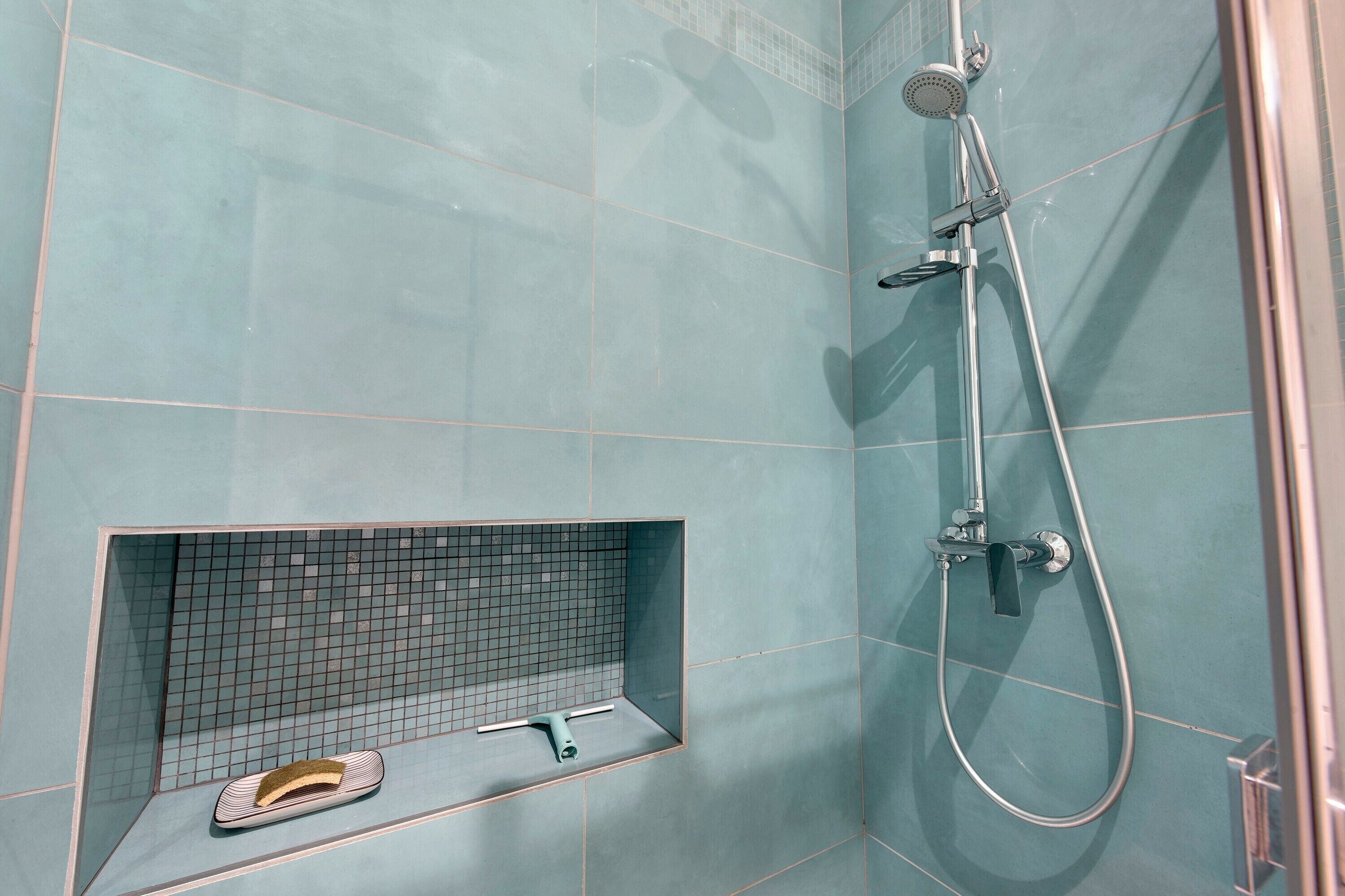 A bathroom with a shower, a tiled wall, and a shelf with a pair of slippers.