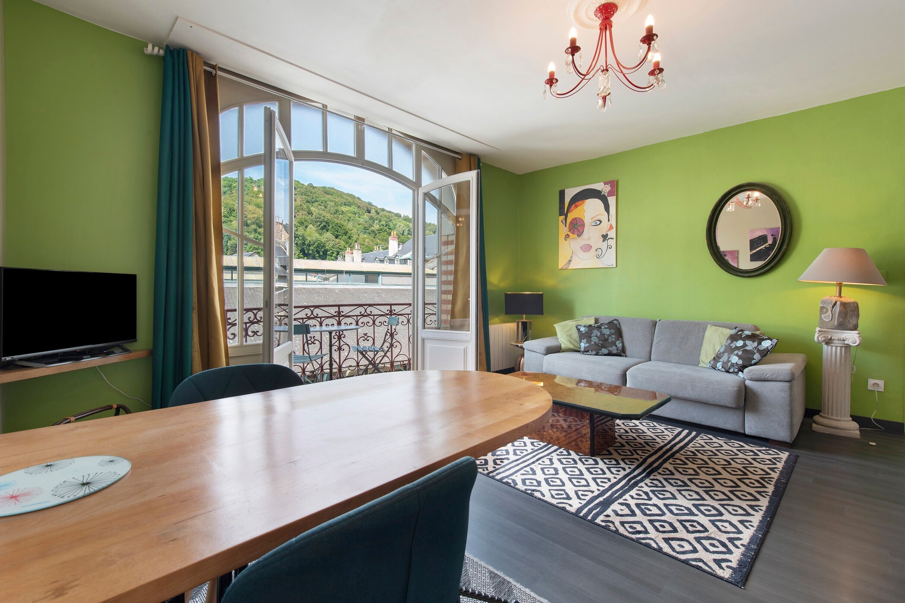 Een woonkamer met een bank, een eettafel, een televisie en een balkon met uitzicht op een heuvel.