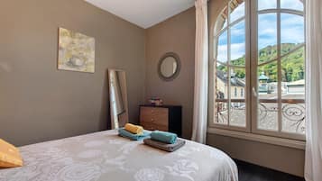 A bedroom with a bed, a dresser, a mirror, and a window with a view of buildings and hills.