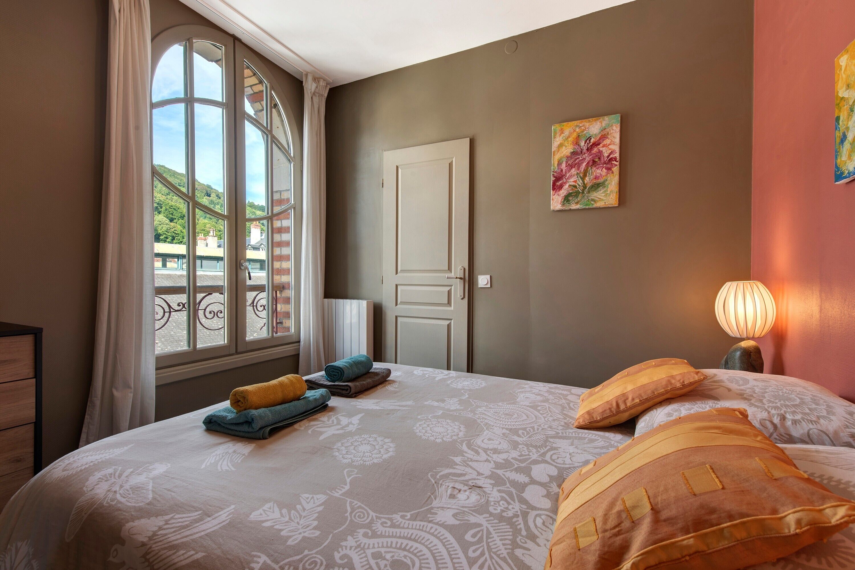 A bedroom with a large window, a bed with patterned bedding, a bedside lamp, and a door.