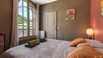 A bedroom with a large window, a bed with patterned bedding, a bedside lamp, and a door.