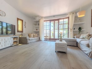 Uma sala de estar espaçosa com uma televisão de tela grande, um armário branco com gavetas, um sofá, uma mesa de centro e uma janela ampla com cortinas.