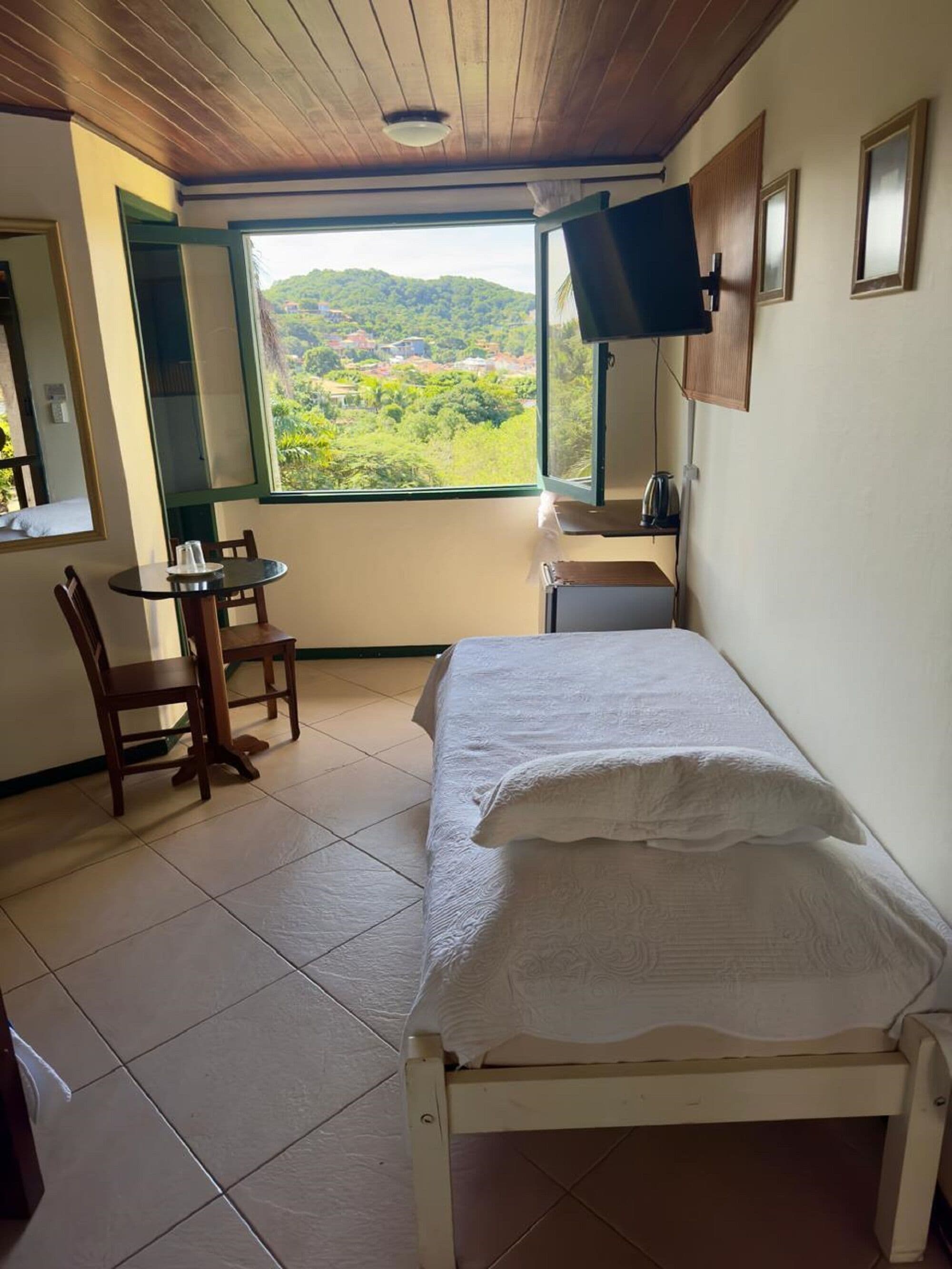 Quarto com cama, cadeira, mesa, janela com vista para o verde e televisão fixada na parede.