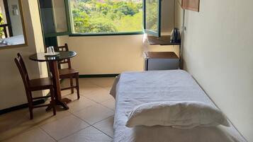Quarto com cama, cadeira, mesa, janela com vista para o verde e televisão fixada na parede.
