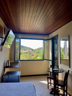 Quarto com cama, mesa com cadeiras, televisão e janela com vista aberta para a paisagem.