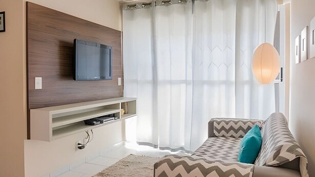 A modern living room with a flat-screen TV mounted on a wooden panel, a sofa with patterned cushions, and a large window with sheer curtains.