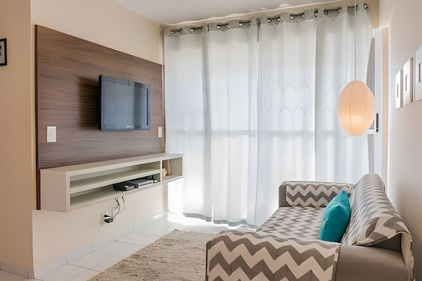 Un salon moderne avec un téléviseur à écran plat fixé sur un panneau en bois, un canapé agrémenté de coussins à motifs et une grande fenêtre ornée de rideaux légers.