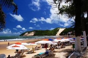 A beach with umbrellas and lounge chairs, a sandy shore, and a hill in the background. - 4let Verano Ponta Negra (Natal)