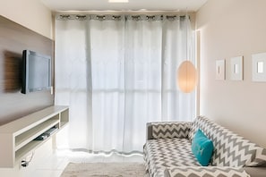 A modern living room with a flat-screen TV mounted on the wall, a sofa with patterned cushions, a large window with sheer curtains, and a pendant light. - 4let Verano Ponta Negra (Natal)