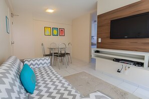 A living room with a sofa, a TV mounted on a wooden panel, a dining table with chairs, and a wall with three framed pictures.