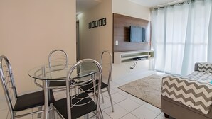 A modern living room with a glass dining table, metal chairs, a sofa, a TV mounted on a wooden panel, and a patterned rug.