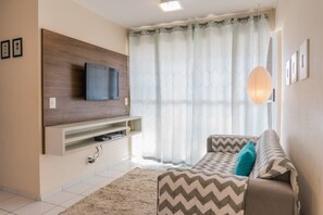 Una sala de estar con un televisor de pantalla plana montado en un panel de madera, un sofá con cojines estampados y un ventanal con cortinas traslúcidas.