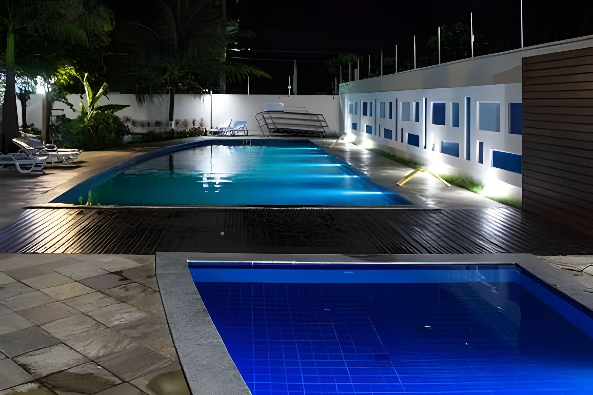 Una piscina con solárium empedrado, sillas de descanso y un edificio con varias ventanas al fondo.