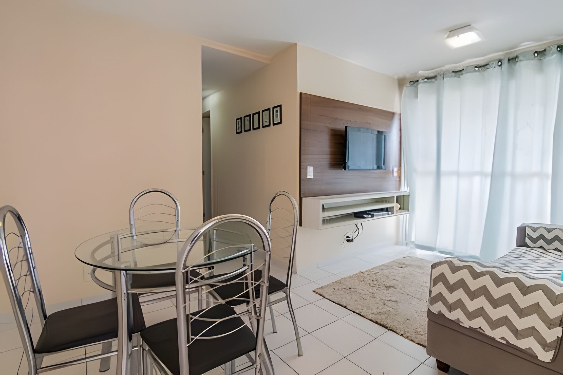 Uma sala de estar moderna com uma mesa de jantar de vidro, cadeiras de metal, um sofá, uma TV fixada em um painel de madeira e um tapete estampado.