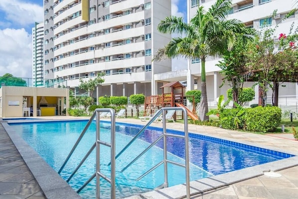 Piscina con escaleras de metal, rodeada de vegetación y un edificio de varios pisos con balcones.