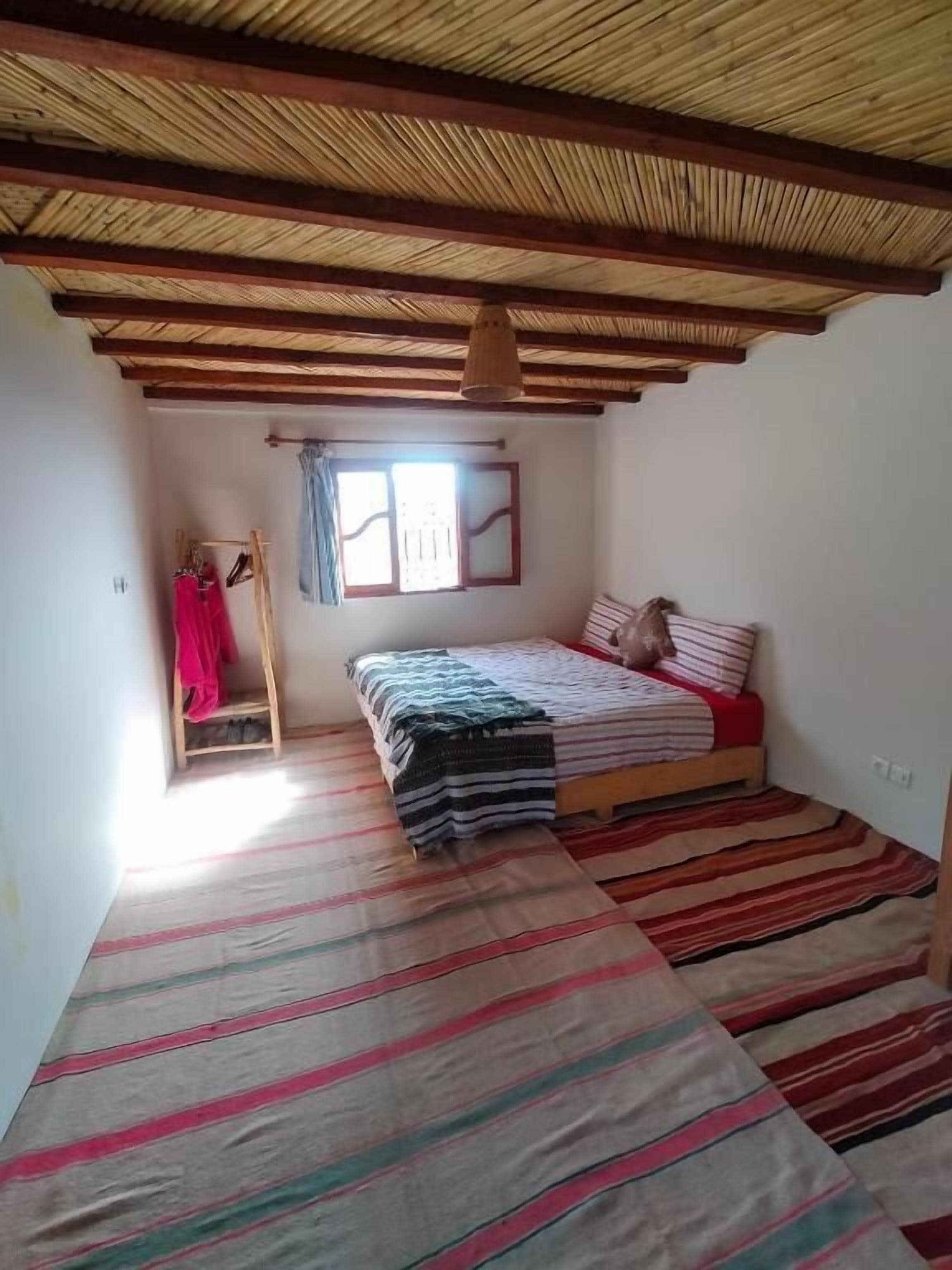 Une pièce avec un lit, une fenêtre avec des rideaux, un plafond en bois et un tapis à rayures.
