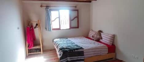 A room with a bed, a window with curtains, a wooden ceiling, and a striped rug.