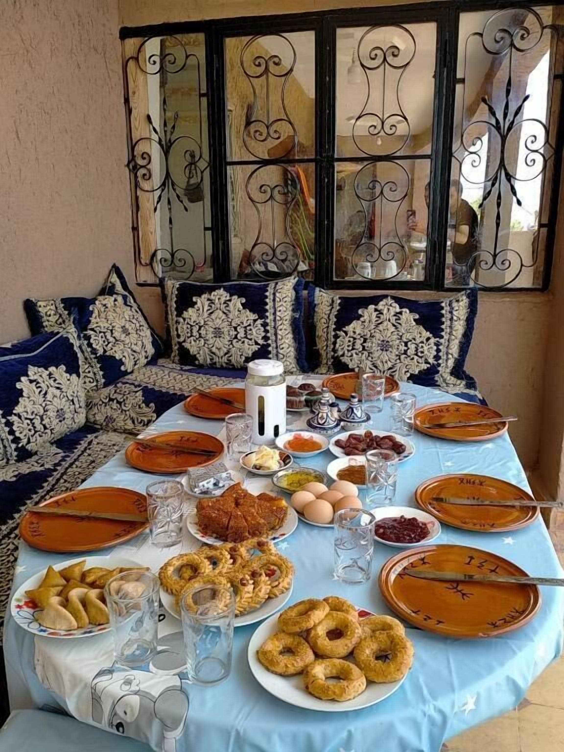Une table à manger dressée avec différents plats, des verres et des couverts, avec une fenêtre décorative et des coussins à motifs en arrière-plan.