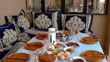 Une table à manger dressée avec différents plats, des verres et des couverts, avec une fenêtre décorative et des coussins à motifs en arrière-plan.