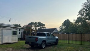 Un camionnette bleu clair était garée dans une cour verdoyante, avec une clôture en métal et un petit abri de jardin en arrière-plan.