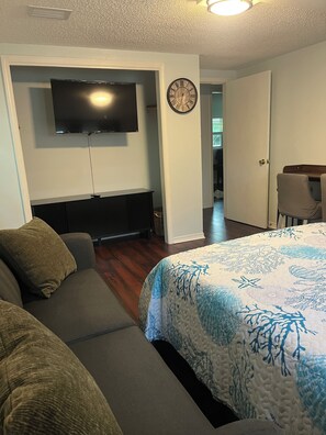Ein Hotelzimmer mit einem Bett, einem Sofa, einem Fernseher, einer Uhr und einer Tür zu einem weiteren Raum.