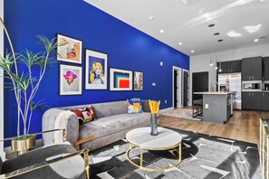 Un salon moderne avec un mur d’accent bleu, un canapé gris, une table basse ronde et un coin cuisine doté de meubles sombres et d’un réfrigérateur en acier inoxydable.