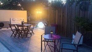 Un patio al aire libre con mesa y sillas de madera, una pequeña mesa redonda con lámpara y un cartel al fondo.