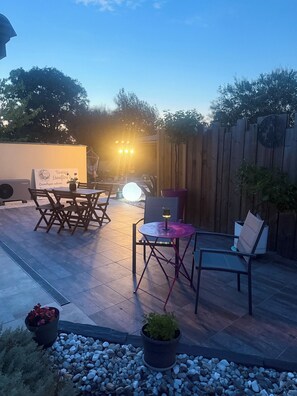 An outdoor patio area with a wooden table and chairs, a small round table with a lamp, and a sign in the background. - Between Bordeaux and Saint-Émilion – Charming Studio with a Pool (La Lande-de-Fronsac)